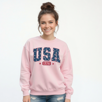 Smiling woman in a light pink crewneck sweatshirt with a navy USA graphic filled with white stars and red 1776 lettering