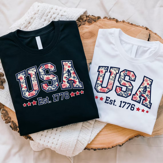 Flat lay of folded black and white USA floral shirts – Est. 1776 patriotic summer T-shirt set.


