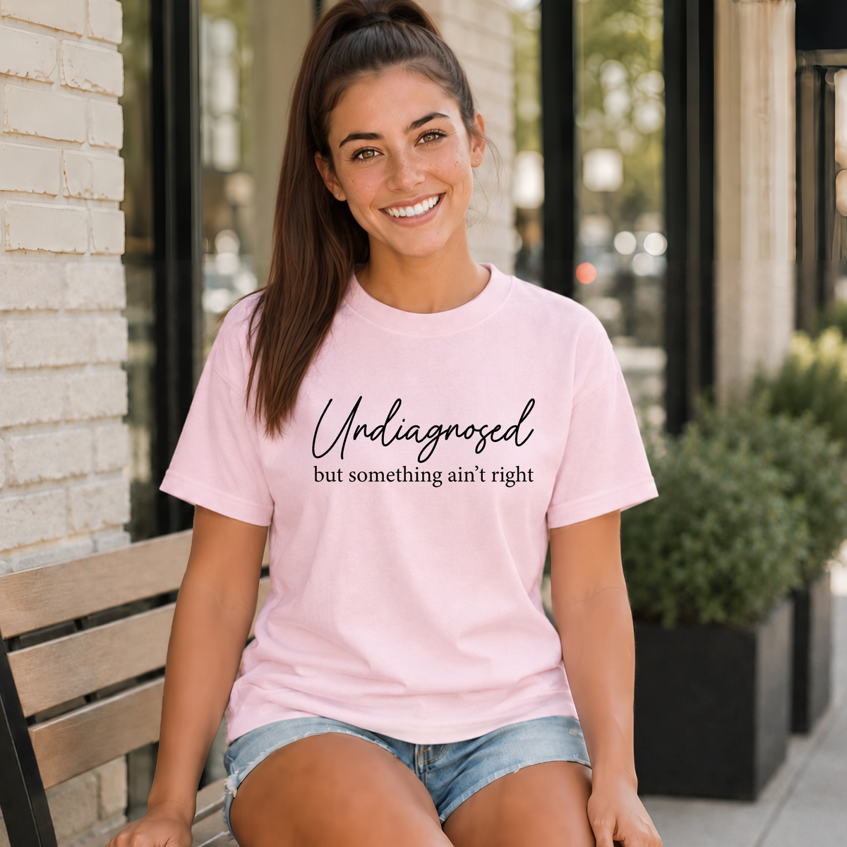 Woman seated on a bench wearing a light pink T-shirt with black Undiagnosed but something ain’t right graphic text.