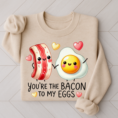Flat lay of a sand-colored sweatshirt with a cute bacon and egg holding hands and colorful hearts, captioned “You're the Bacon to My Eggs.”