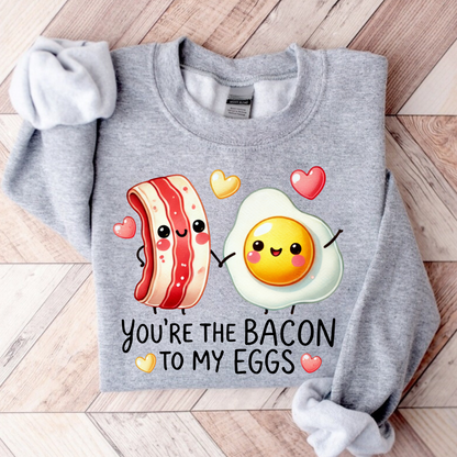 Folded sport gray sweatshirt laid on wooden background, printed with a smiling bacon and egg graphic and “You're the Bacon to My Eggs” text.