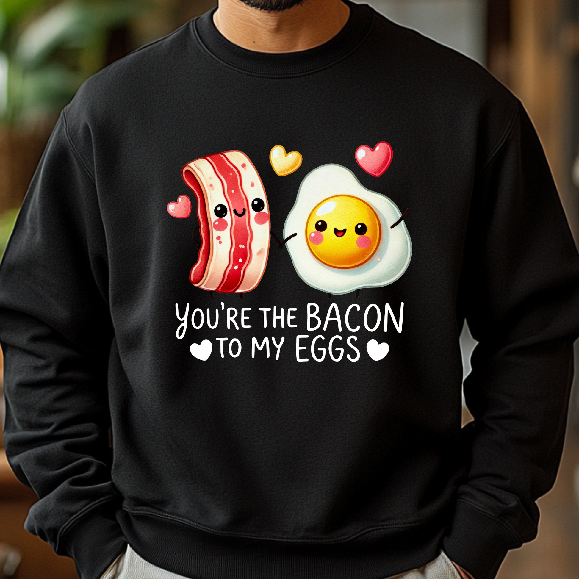 Man wearing a black sweatshirt featuring a cute cartoon bacon and egg holding hands with hearts, and the text “You're the Bacon to My Eggs.”