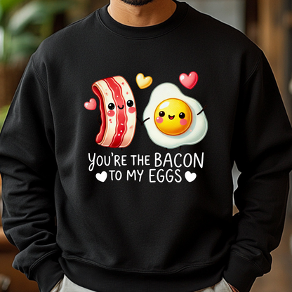 Man wearing a black sweatshirt featuring a cute cartoon bacon and egg holding hands with hearts, and the text “You're the Bacon to My Eggs.”
