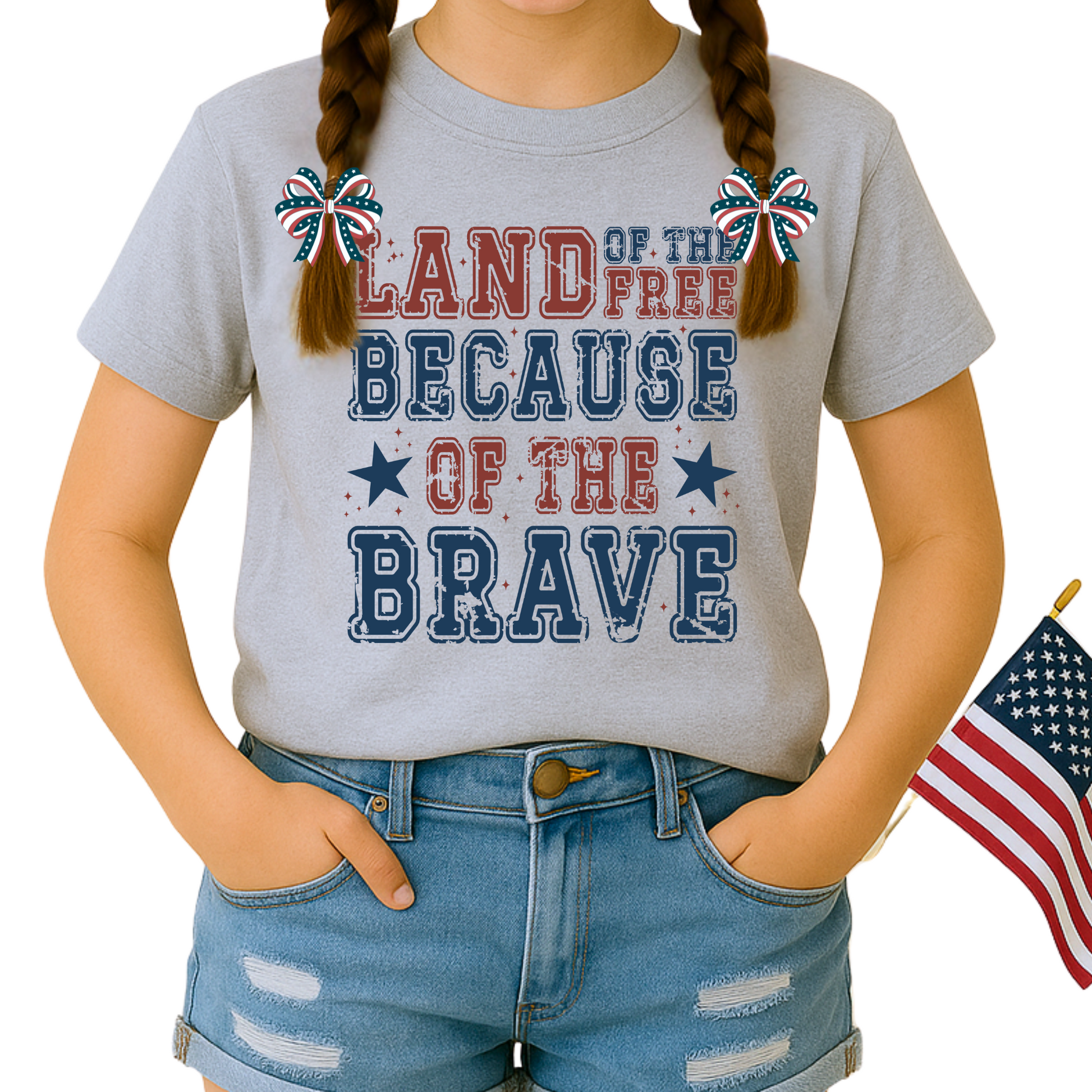 Smiling girl in Sport Gray  "Land of the Free Because of the Brave" shirt, styled with American flag hair bows and denim shorts