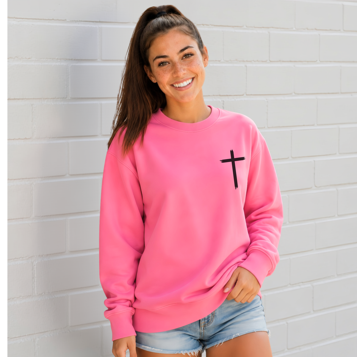 Woman wearing a bright pink crewneck sweatshirt with a black cross on the front chest.