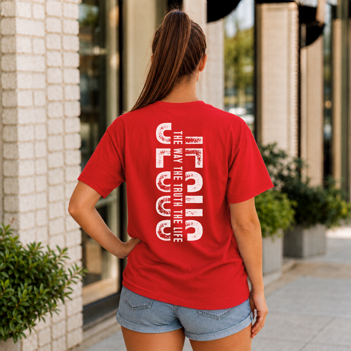 Woman shown from behind wearing a red T-shirt with vertical white JESUS lettering and “The Way, The Truth, The Life” text.