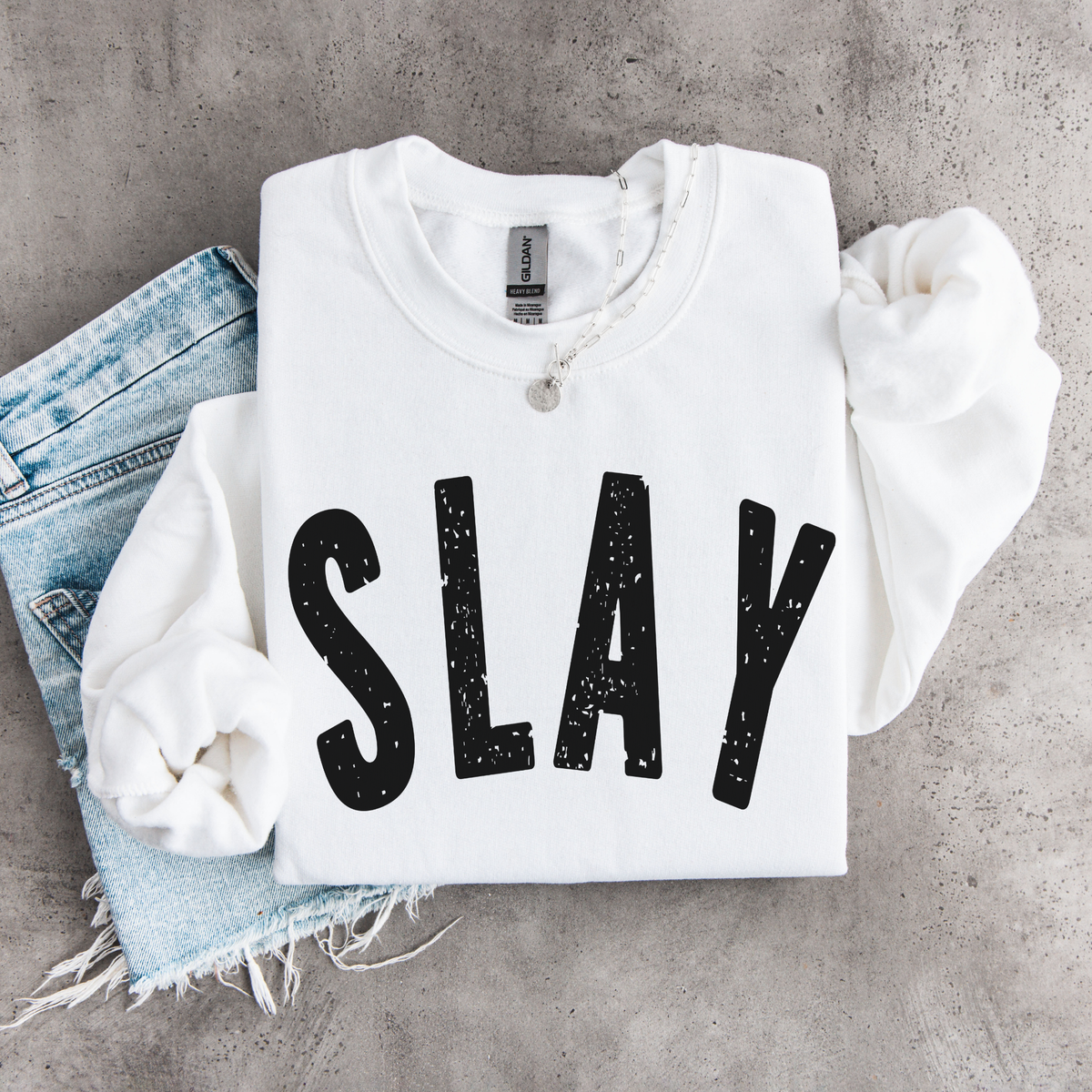 White SLAY crewneck sweatshirt flat lay with bold black distressed lettering, styled with denim, necklace, and matching scrunchies.