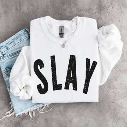 White SLAY crewneck sweatshirt flat lay with bold black distressed lettering, styled with denim, necklace, and matching scrunchies.