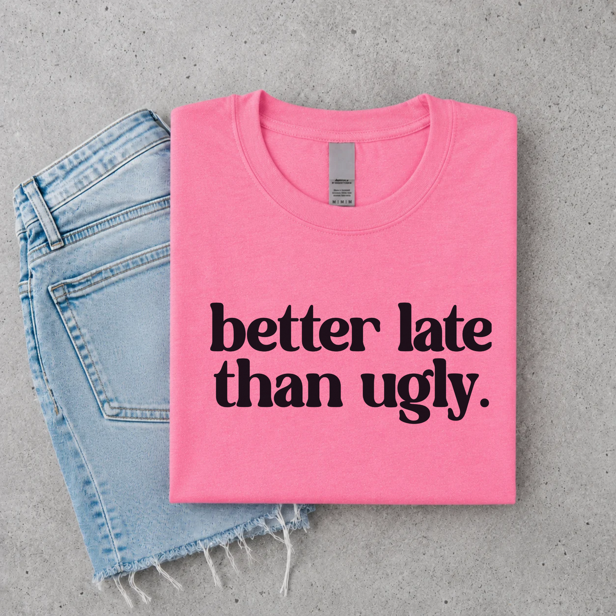 Bright pink Better Late Than Ugly youth T-shirt flat lay with black lettering, styled with denim.