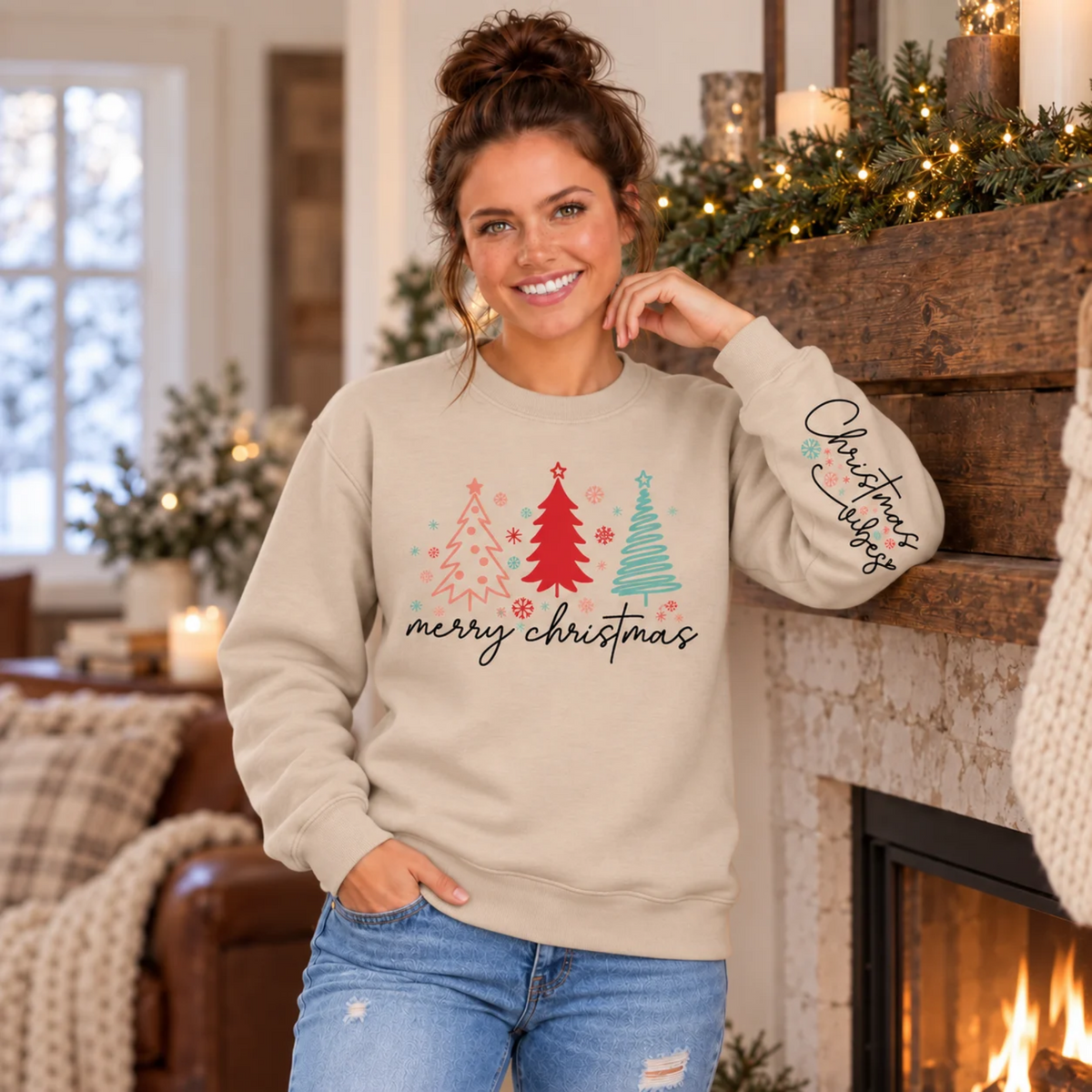 Woman wearing a sand Merry Christmas tree crewneck sweatshirt with Christmas Vibes sleeve print beside a decorated fireplace with warm holiday greenery and lights.