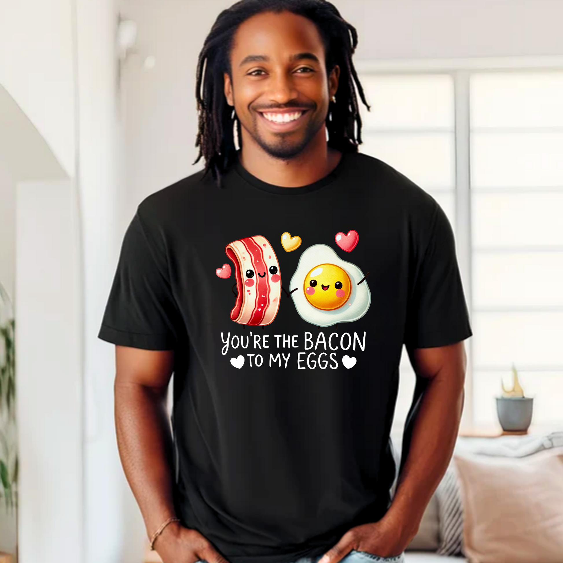 Smiling man wearing a black T-shirt with kawaii-style bacon and egg holding hands, surrounded by colorful hearts. Design says 'You're the bacon to my eggs