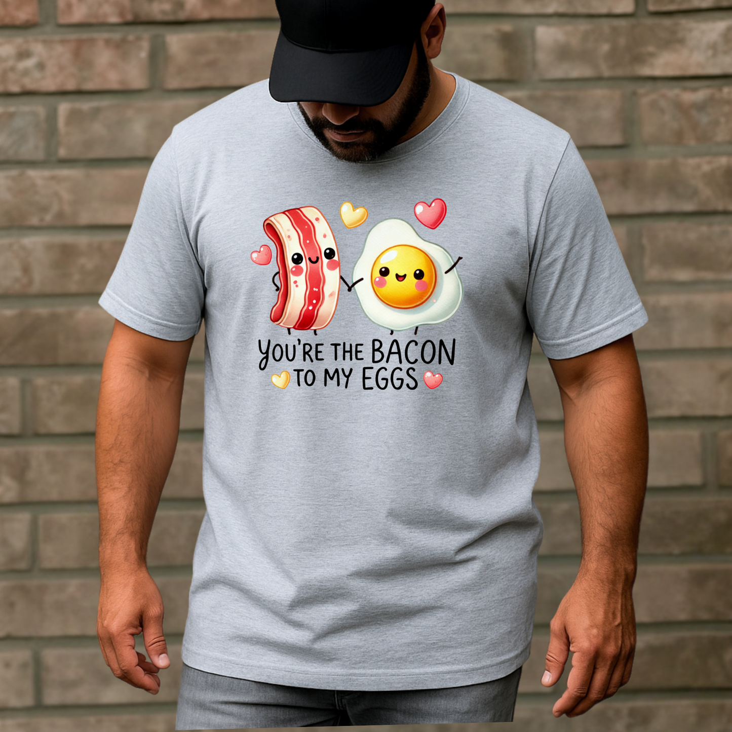 Man in a gray T-shirt featuring adorable smiling bacon and egg characters holding hands with hearts above them. Text says 'You're the bacon to my eggs.'"