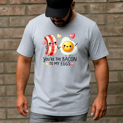 Man in a gray T-shirt featuring adorable smiling bacon and egg characters holding hands with hearts above them. Text says 'You're the bacon to my eggs.'"
