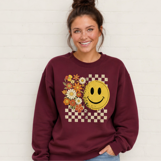 Woman wearing a maroon crewneck sweatshirt with a retro yellow smiley face, fall flowers, and checkerboard graphic.