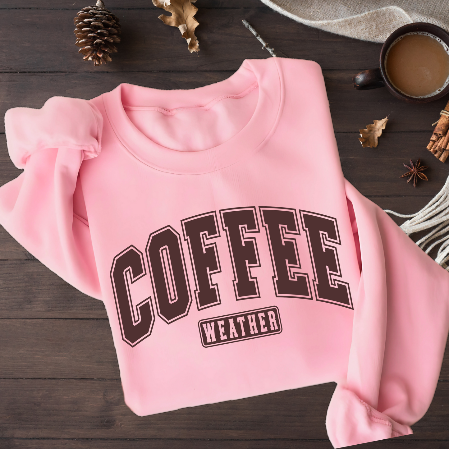 Pink sweatshirt with 'COFFEE WEATHER' text on a wooden surface with coffee and decorative items.