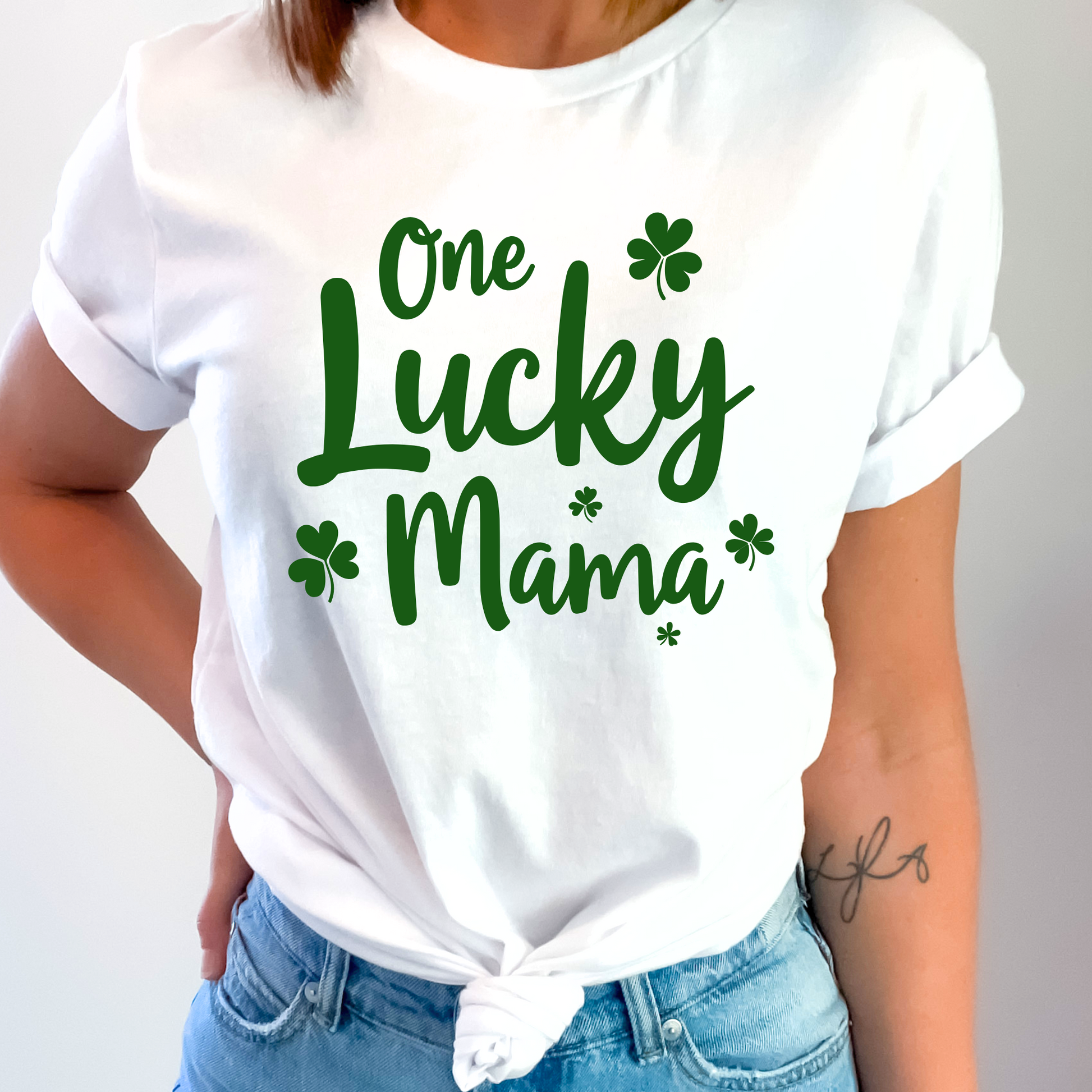 A stylish white t-shirt with "One Lucky Mama" in green script, decorated with shamrocks. Perfect for moms celebrating St. Patrick’s Day in a cute and festive way.