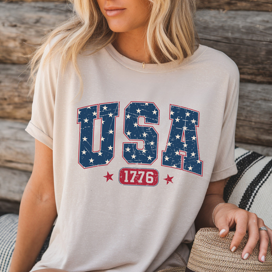 USA 1776 graphic T-shirt in sand color, patriotic tee styled for summer by woman sitting on wooden bench