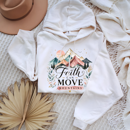 White hoodie with 'Faith Can Move Mountains' design on a textured surface with a hat and leaves.