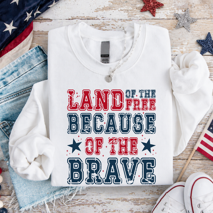 White sweatshirt with 'Land of the Free Because of the Brave' text on a wooden surface with American flags and denim jeans.