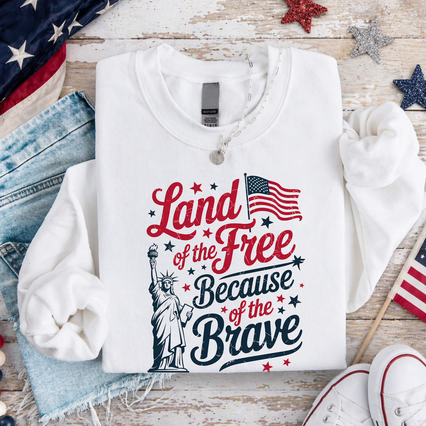 White sweatshirt with 'Land of the Free Because of the Brave' text and American flag design on a wooden surface.