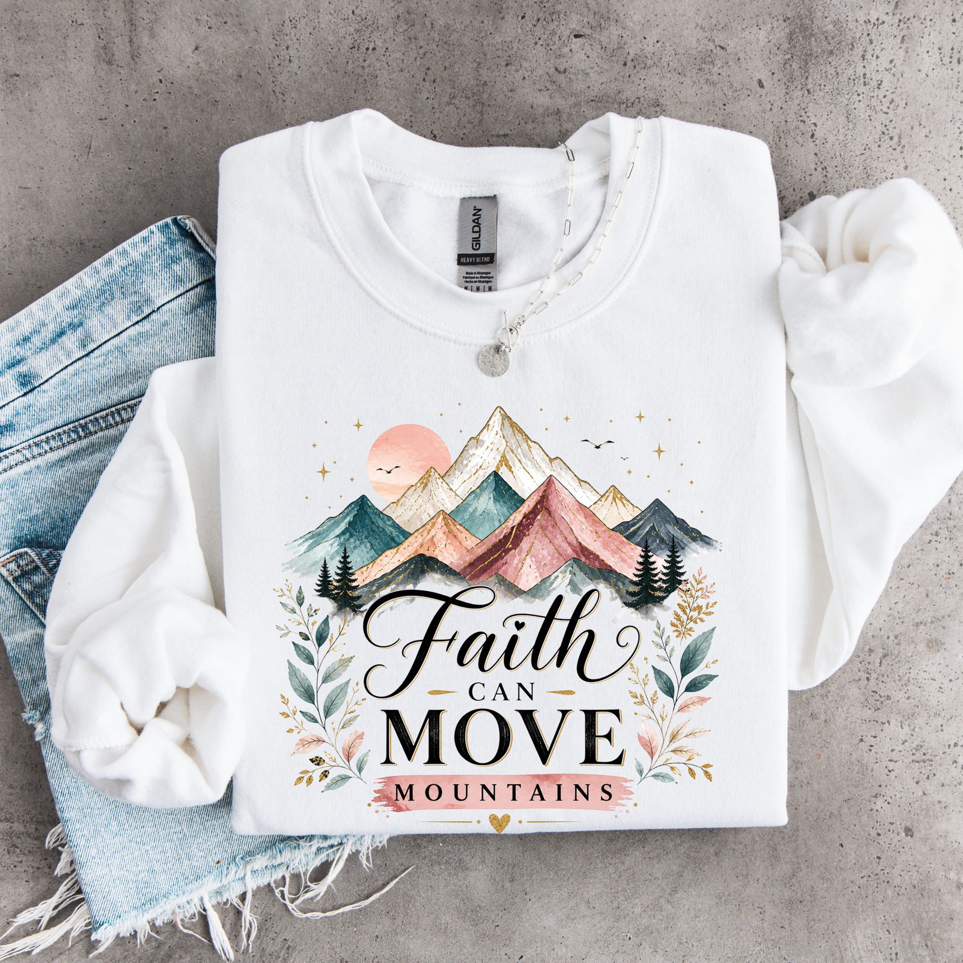 White sweatshirt with mountain design and 'Faith Can Move Mountains' text on a concrete surface.