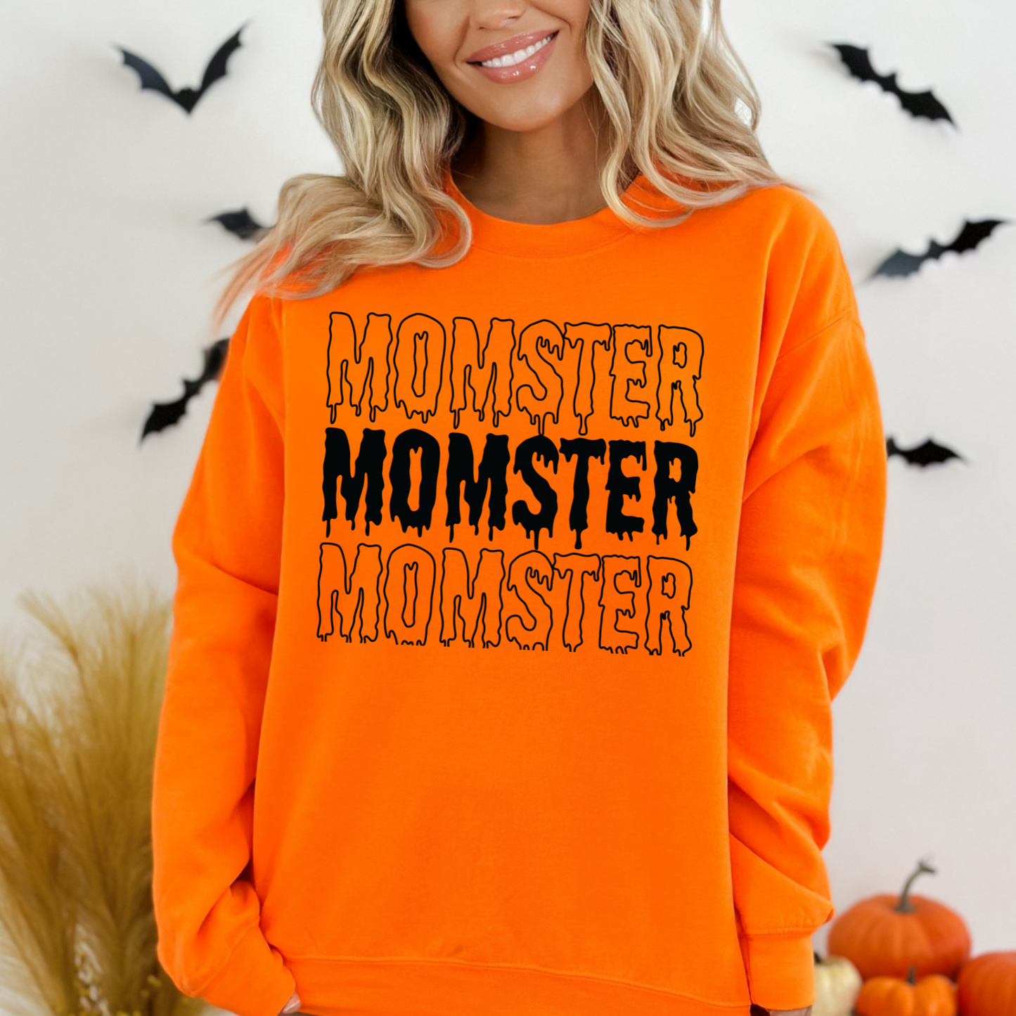 Young mom wearing an orange sweatshirt with 'MONSTER' text, Halloween-themed background with pumpkins and bats.