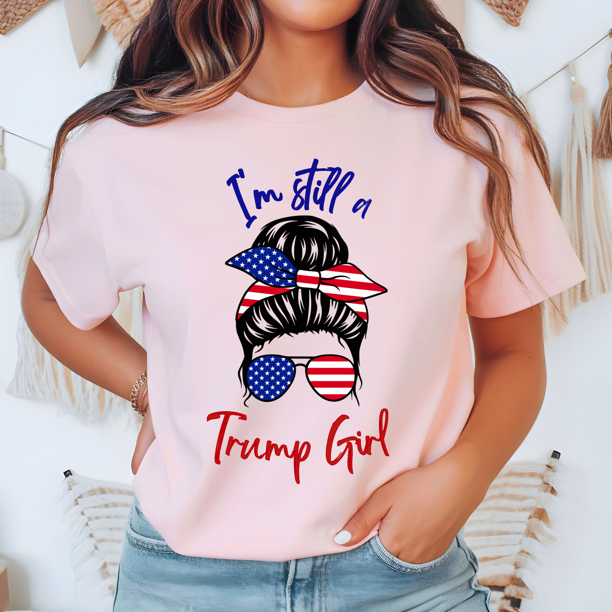 Woman wearing pink “I’m Still a Trump Girl” patriotic T-shirt with USA flag bun and sunglasses design.