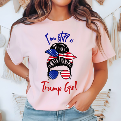 Woman wearing pink “I’m Still a Trump Girl” patriotic T-shirt with USA flag bun and sunglasses design.