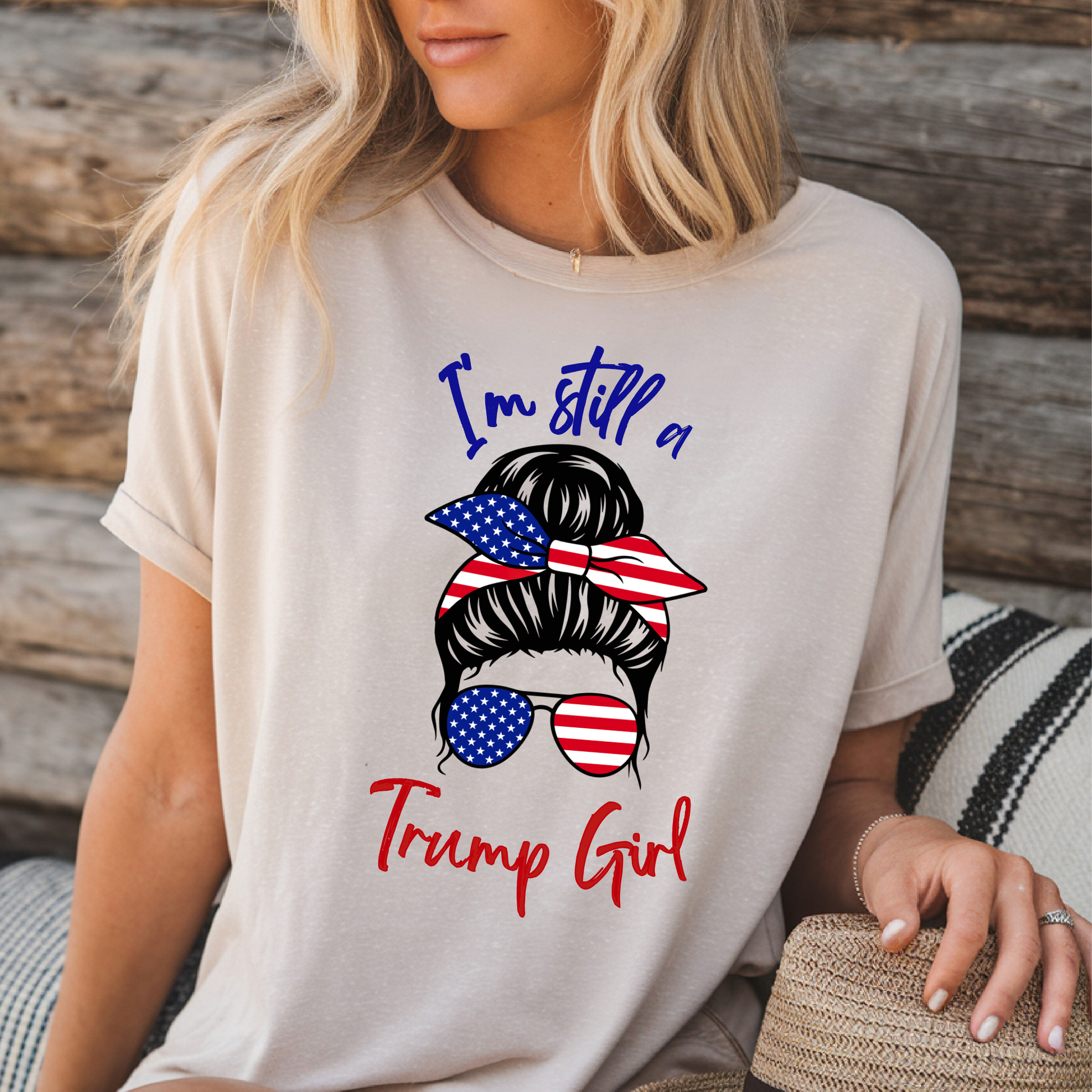 Woman wearing Sand “I’m Still a Trump Girl” patriotic T-shirt with USA flag bun and sunglasses design.