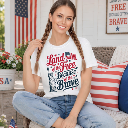Woman wearing a 'Land of the Free Because of the Brave' t-shirt sitting outdoors with an American flag in the background.