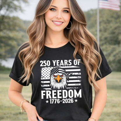 Woman wearing a black t-shirt with '250 Years of Freedom' design, standing outdoors with trees and an American flag in the background.