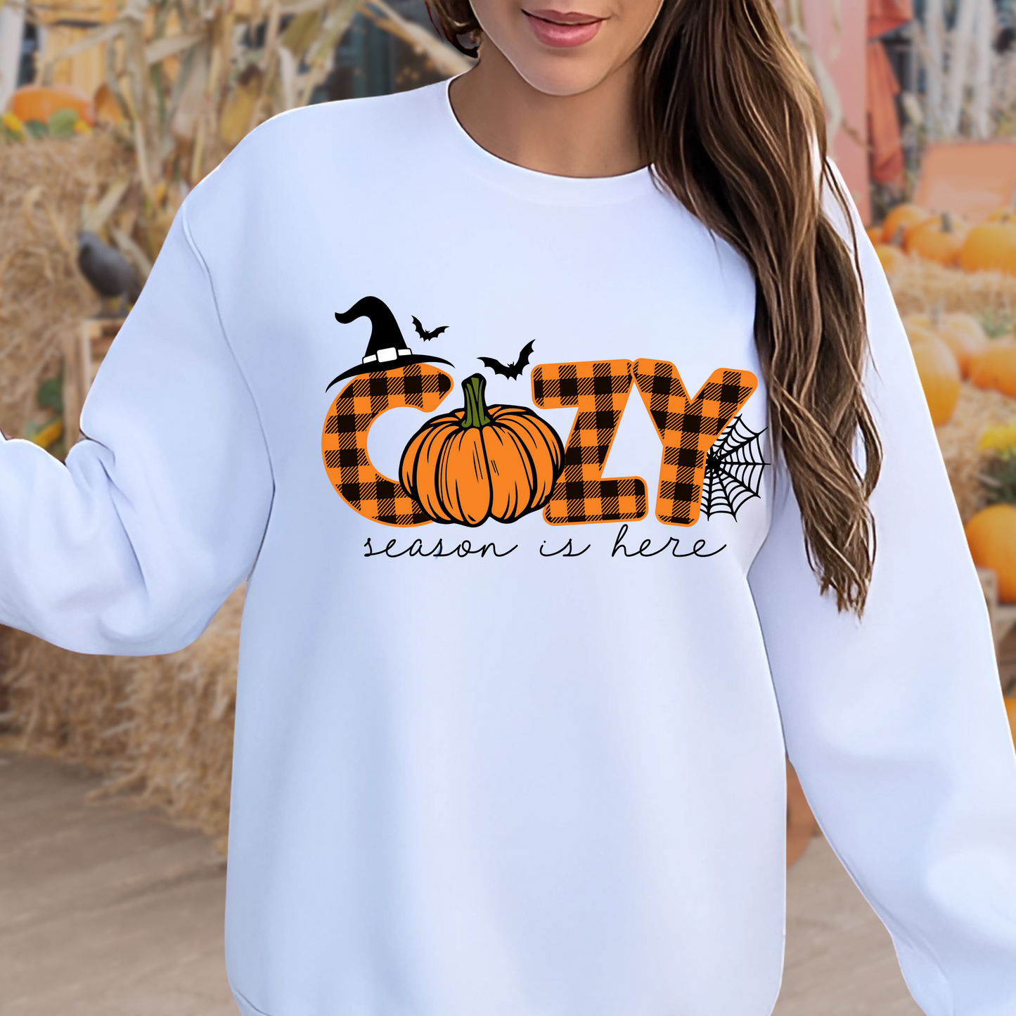 Woman wearing a white fall sweatshirt featuring the phrase “Cozy Season Is Here” with a buffalo plaid pumpkin, witch hat, bats, and spiderweb – set against a pumpkin patch background.
