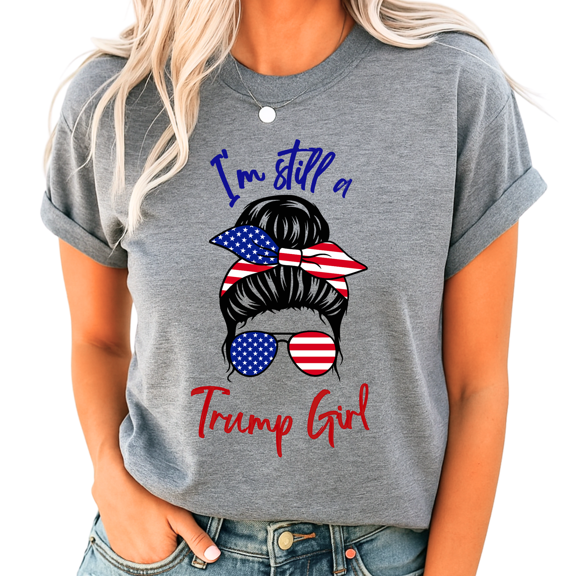 Woman wearing gray “I’m Still a Trump Girl” patriotic T-shirt with USA flag bun and sunglasses design.