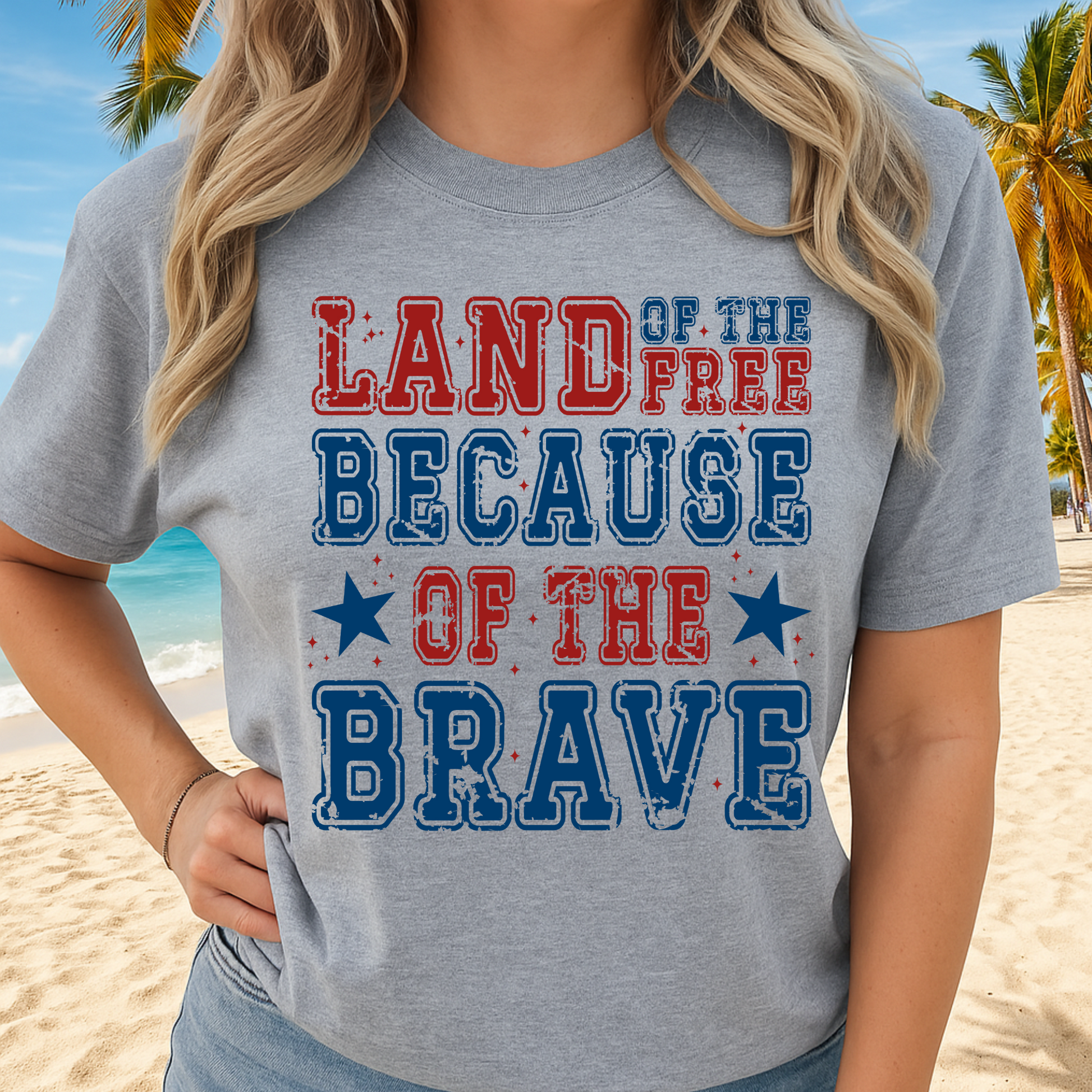 Woman wearing gray t-shirt with “Land of the Free Because of the Brave” design, standing on the beach

