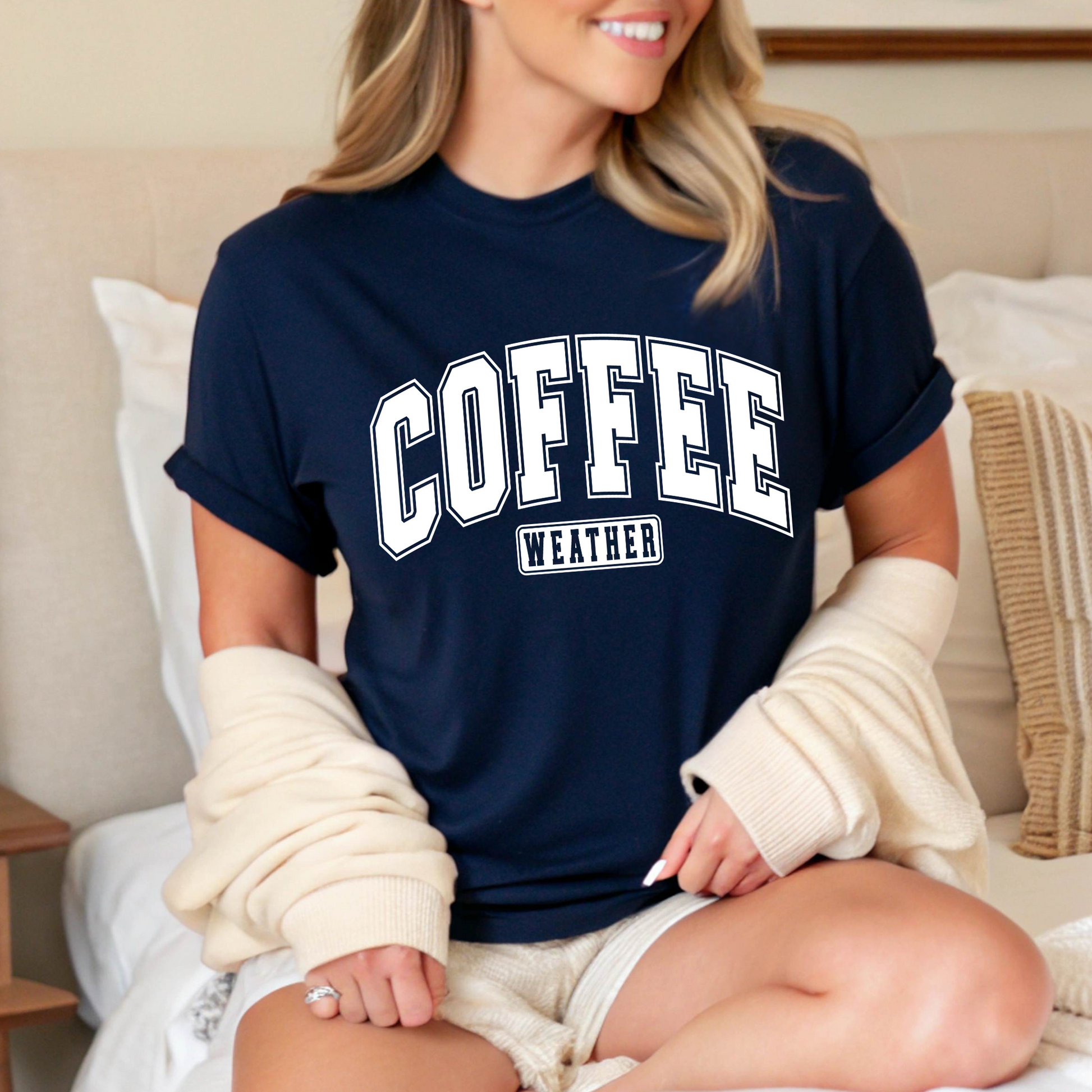 Woman wearing navy "Coffee Weather" shirt sitting on bed, cozy fall look