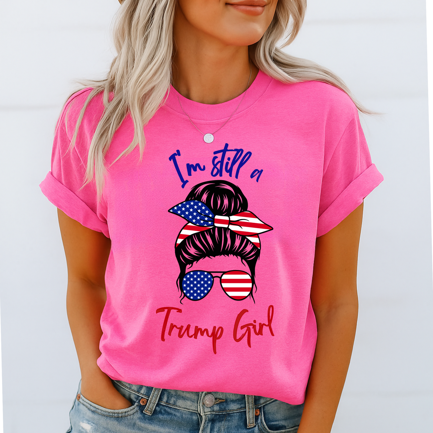 Woman wearing pink “I’m Still a Trump Girl” patriotic T-shirt with USA flag bun and sunglasses design.