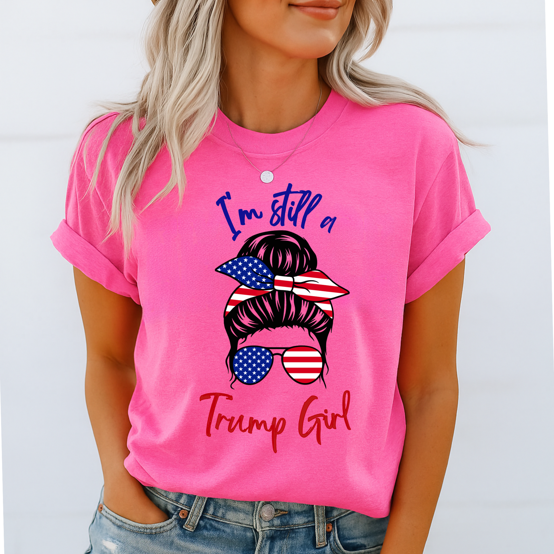 Woman wearing pink “I’m Still a Trump Girl” patriotic T-shirt with USA flag bun and sunglasses design.