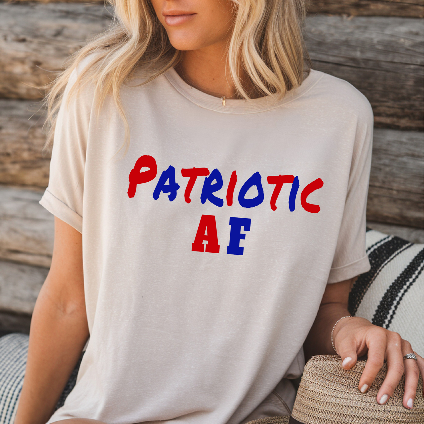Woman wearing tan “Patriotic AF” graphic t-shirt seated against rustic wood