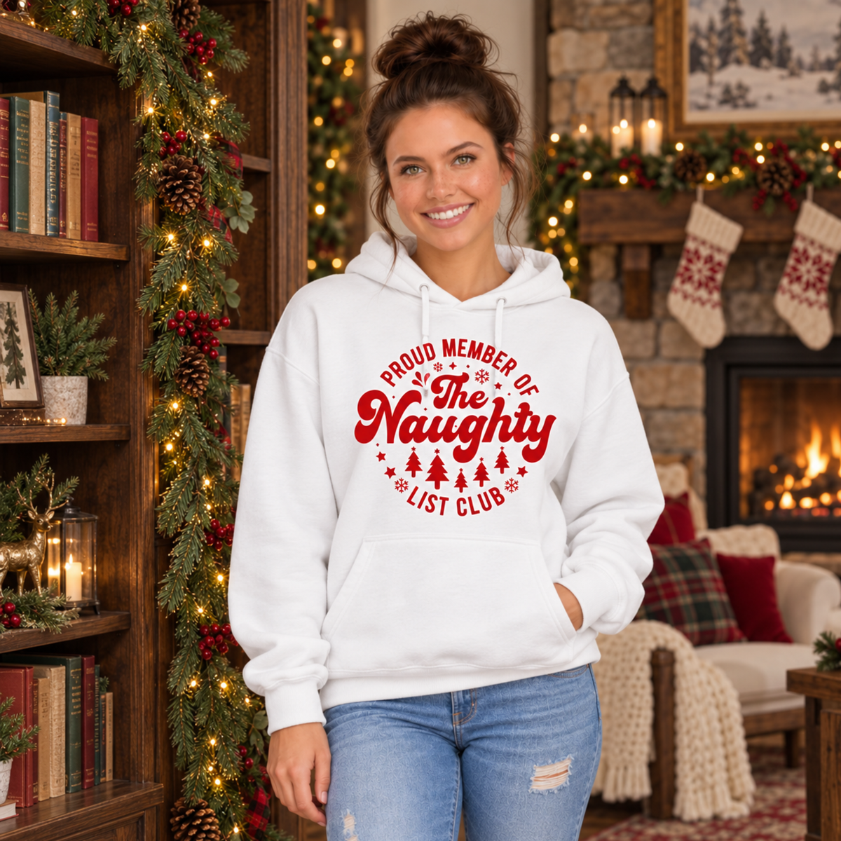 Woman wearing a white hoodie with a red Proud Member of the Naughty List Club design in a cozy Christmas room with a fireplace.
