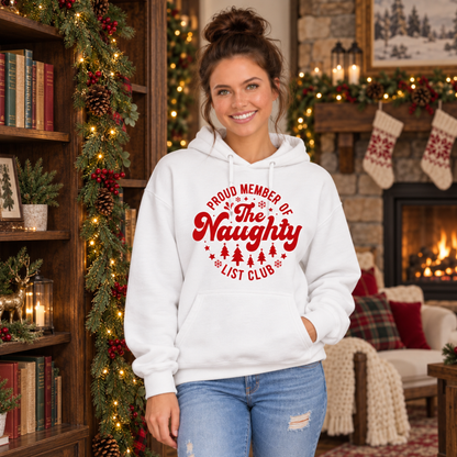 Woman wearing a white hoodie with a red Proud Member of the Naughty List Club design in a cozy Christmas room with a fireplace.