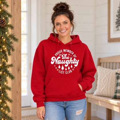 Woman wearing a red hoodie with a white Proud Member of the Naughty List Club design beside lit Christmas garland.