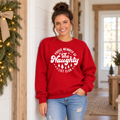 Woman wearing a red crewneck sweatshirt with a white Proud Member of the Naughty List Club design in a simple festive hallway.