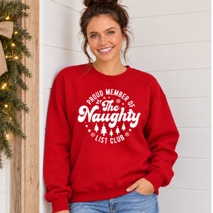 Woman wearing a red crewneck sweatshirt with a white Proud Member of the Naughty List Club design beside simple Christmas greenery.