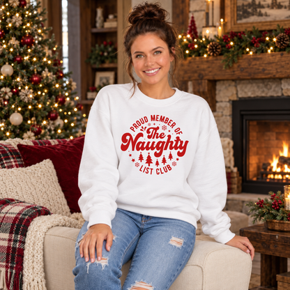 Woman wearing a white crewneck sweatshirt with a red Proud Member of the Naughty List Club design while seated by a Christmas tree and fireplace.