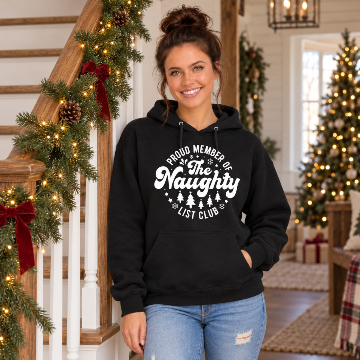 Woman wearing a black hoodie with a white Proud Member of the Naughty List Club design beside a garland-wrapped staircase.