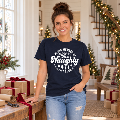 Woman wearing a navy T-shirt with a white Proud Member of the Naughty List Club design beside a holiday gift-wrapping table.