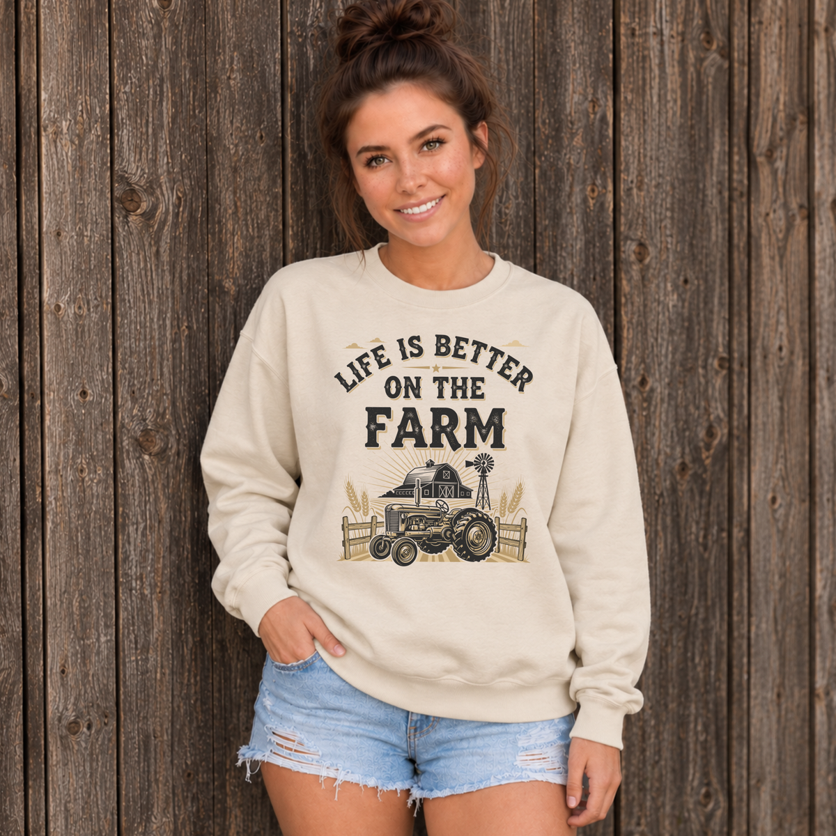 Woman wearing a sand Life Is Better on the Farm sweatshirt against a dark wooden barn wall.