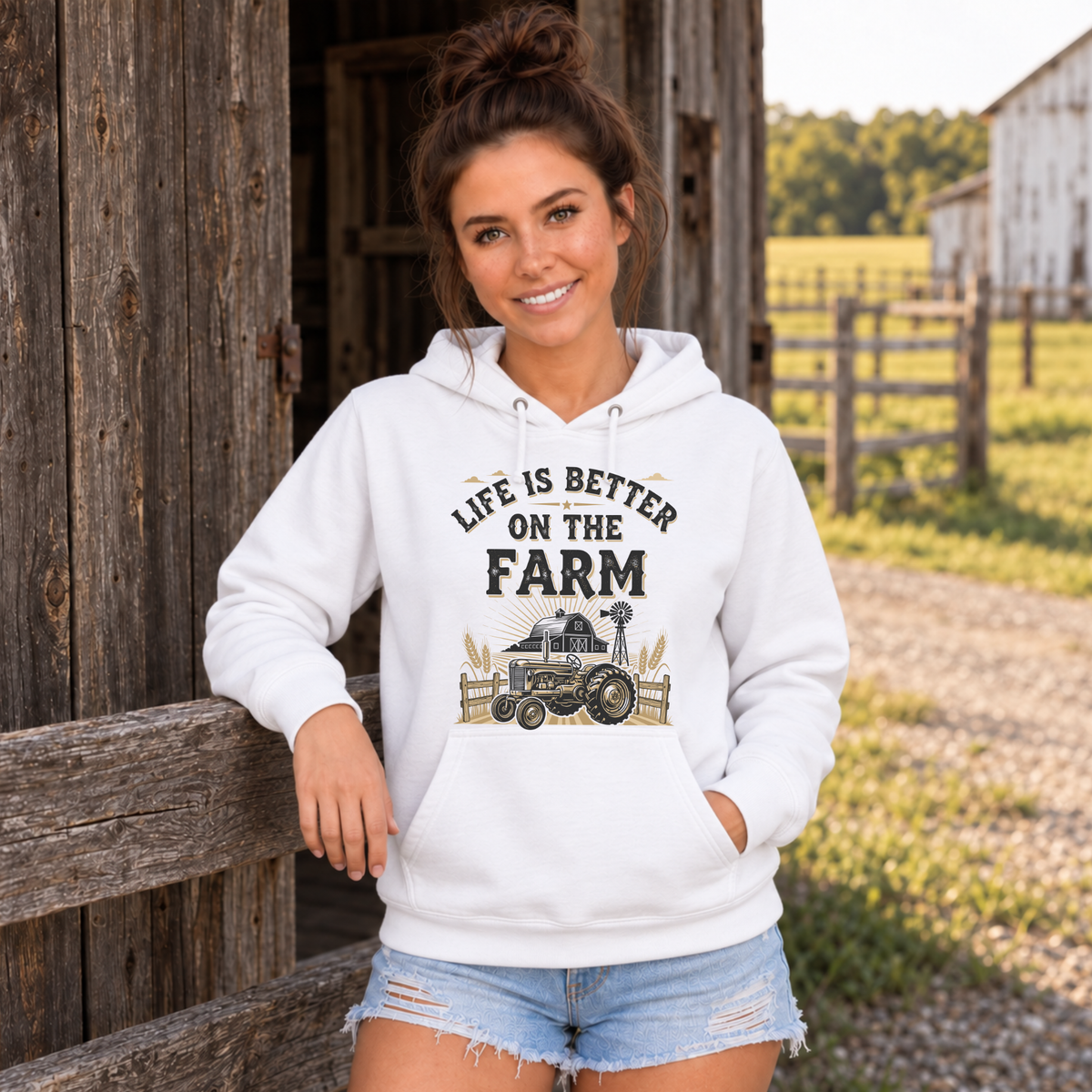 Woman wearing a white Life Is Better on the Farm hoodie beside a rustic barn and fenced field.