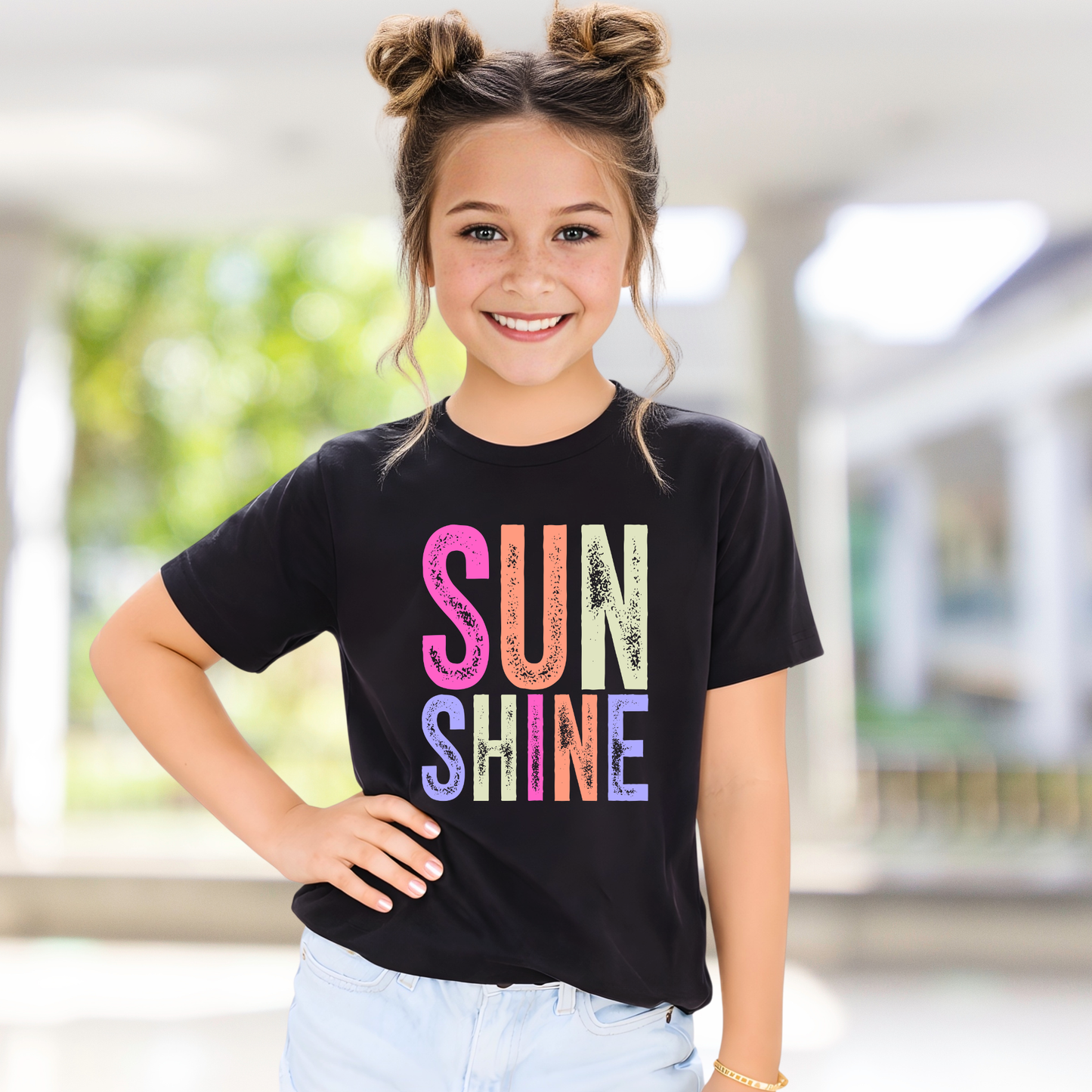 Young girl wearing a black t-shirt with 'SUN SHINE' in colorful letters, standing indoors.