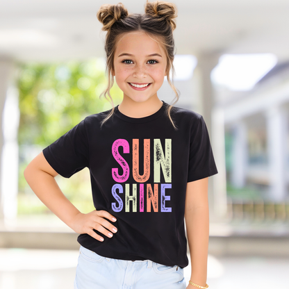 Young girl wearing a black t-shirt with 'SUN SHINE' in colorful letters, standing indoors.